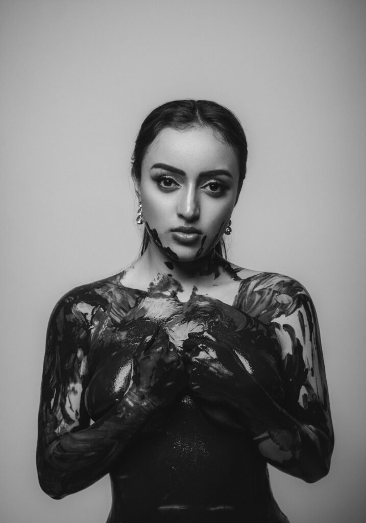 Retrato de Mujer cubierta de pintura negra. Tiene un fondo blanco y la fotografía es a blanco y negro.